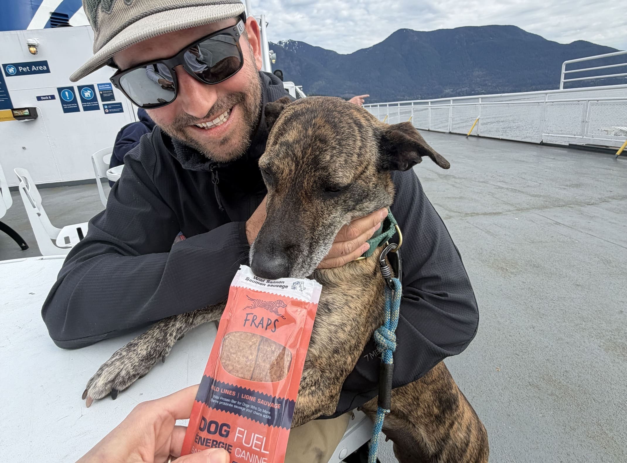 Alma the adventure dog being offered a FRAPS Wild Salmon bar on the BC Ferries pet deck, Howe Sound mountains in the background